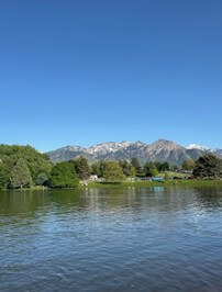 View of a l ake with the Wasatch mountaion range in the background, featuring clear blue skies and lush greenery along the shoreline.